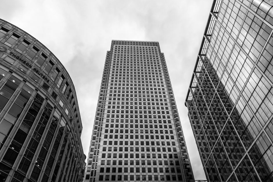 A black and white photograph taken from a low-angle perspective, showcasing three modern high-rise buildings in Canary Wharf. The building on the left features a rounded facade with vertical metal fra